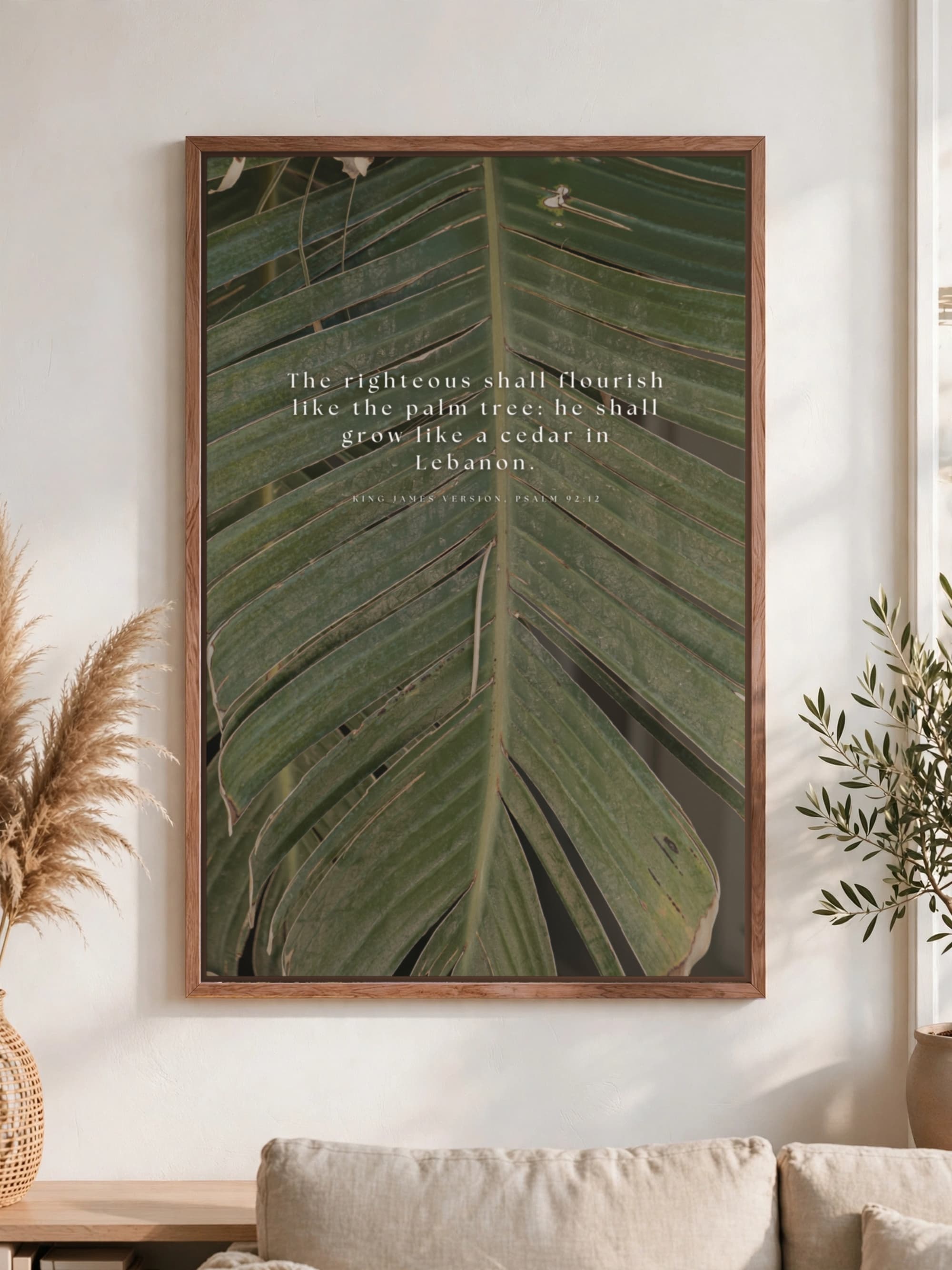 Psalm 92:12 in spaced white serif across a close-up palm frond, pecan frame, hung above a linen sofa.