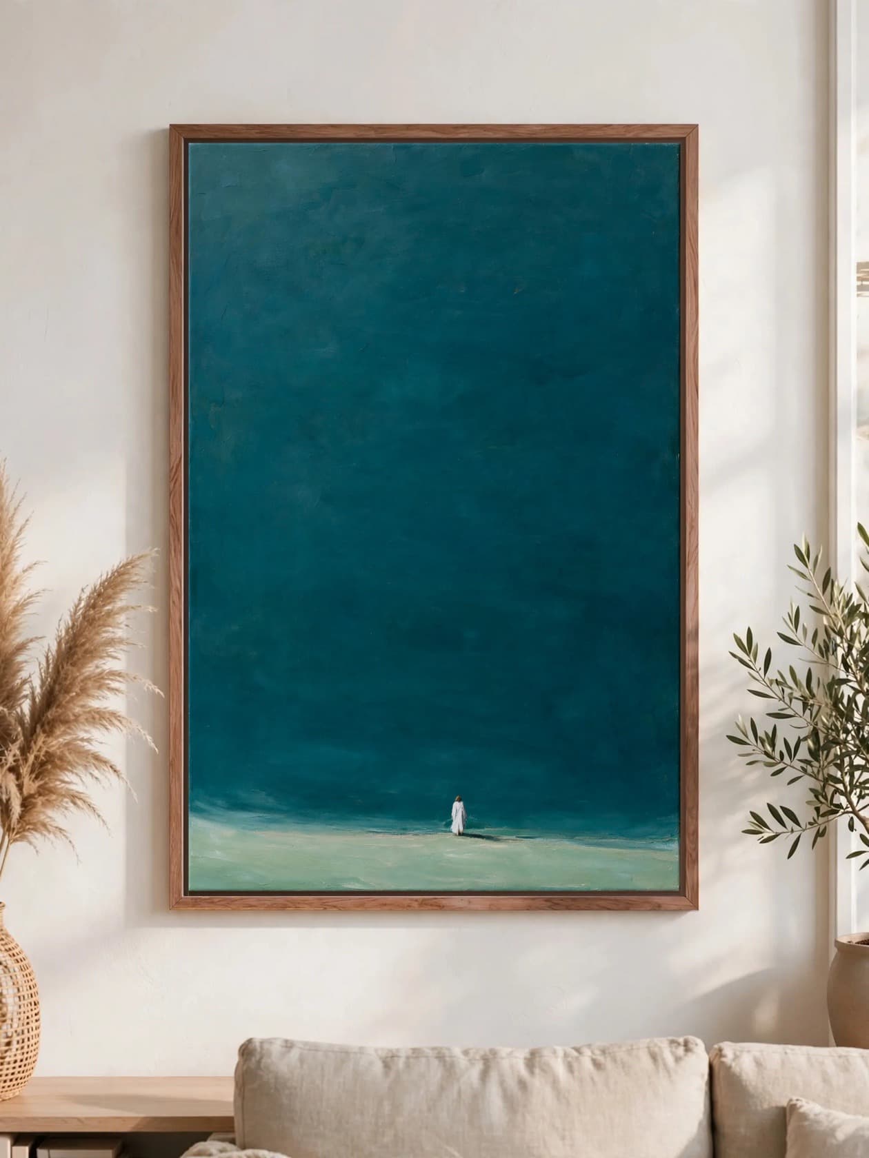 A lone figure in white on a pale green shore beneath a deep teal sky, pecan frame, hung above a linen sofa.